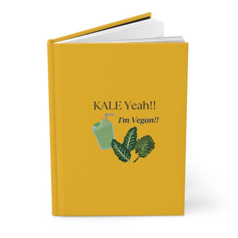 Yellow hardcover journal with the phrase ‘KALE Yeah!! I’m Vegan!!’ printed above illustrations of leafy greens and a green smoothie, placed on a white cloth with dried flowers.