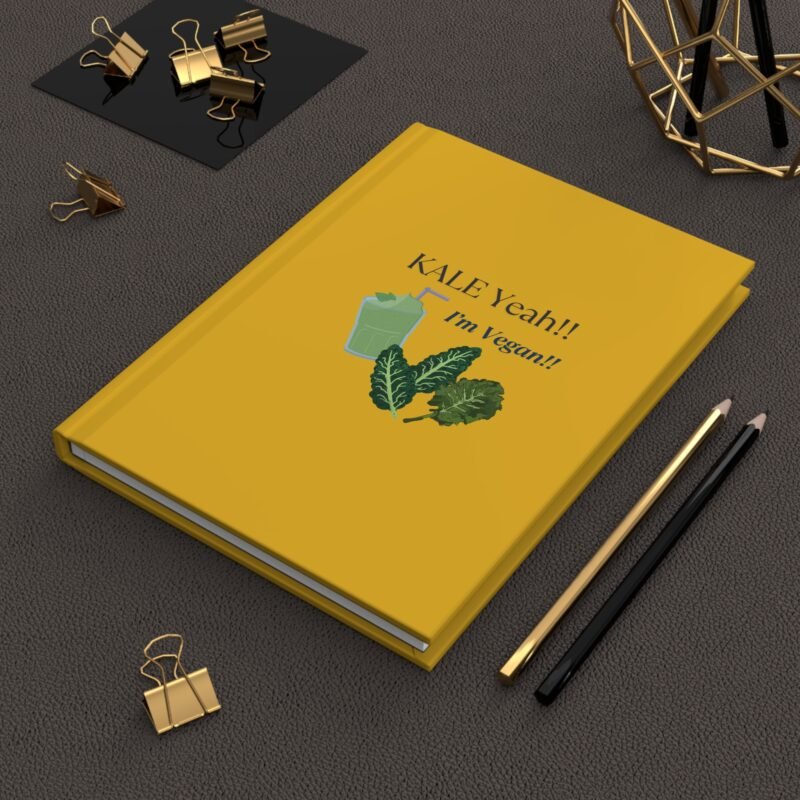 Yellow hardcover journal with the phrase ‘KALE Yeah!! I’m Vegan!!’ printed above illustrations of leafy greens and a green smoothie, placed on a white cloth with dried flowers.