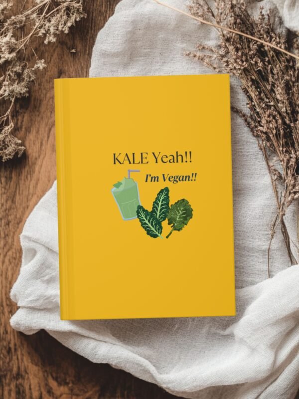Yellow hardcover journal with the phrase ‘KALE Yeah!! I’m Vegan!!’ and illustrations of leafy greens and a green smoothie, styled with dried flowers on a wooden table.