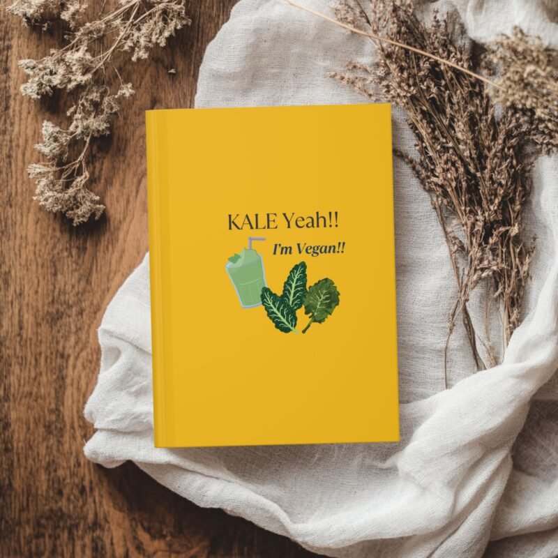 Yellow hardcover journal with the phrase ‘KALE Yeah!! I’m Vegan!!’ and illustrations of leafy greens and a green smoothie, styled with dried flowers on a wooden table.