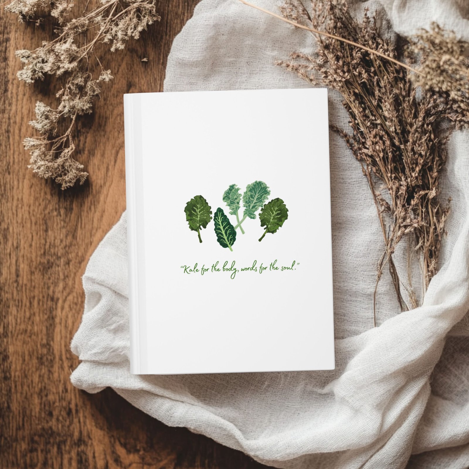 White Matte hardcover journal with the phrase ‘KALE for the body, words for the soul" printed below illustrations of leafy greens kale, placed on a white cloth with dried flowers.